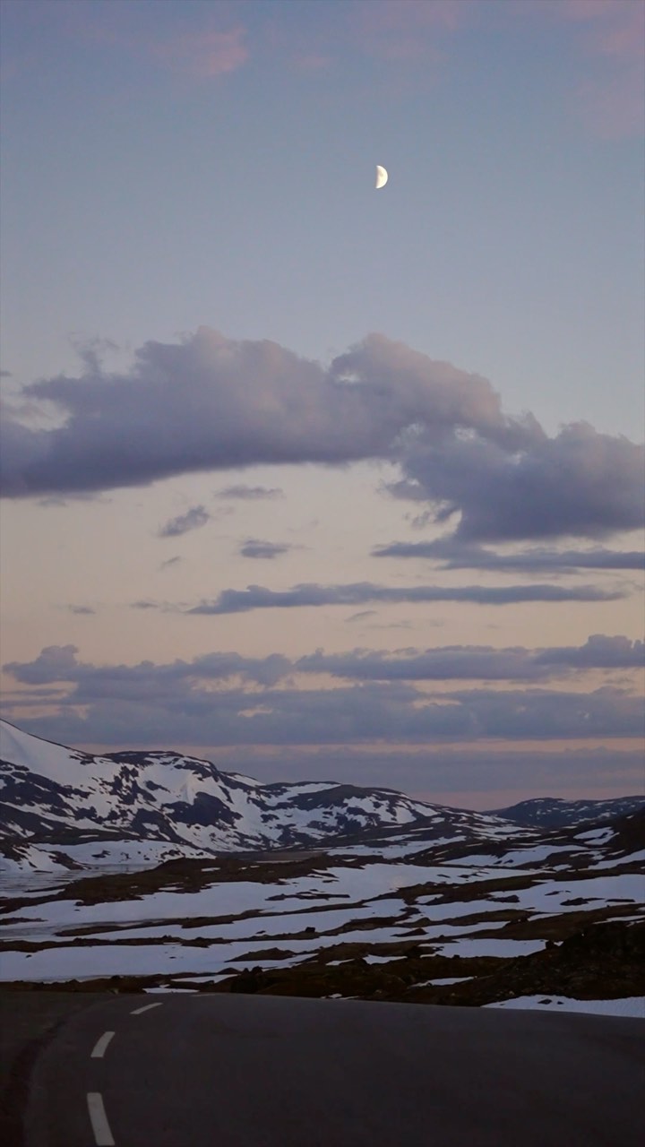 Fjellvei i skumringen med månen over snøkledde fjell